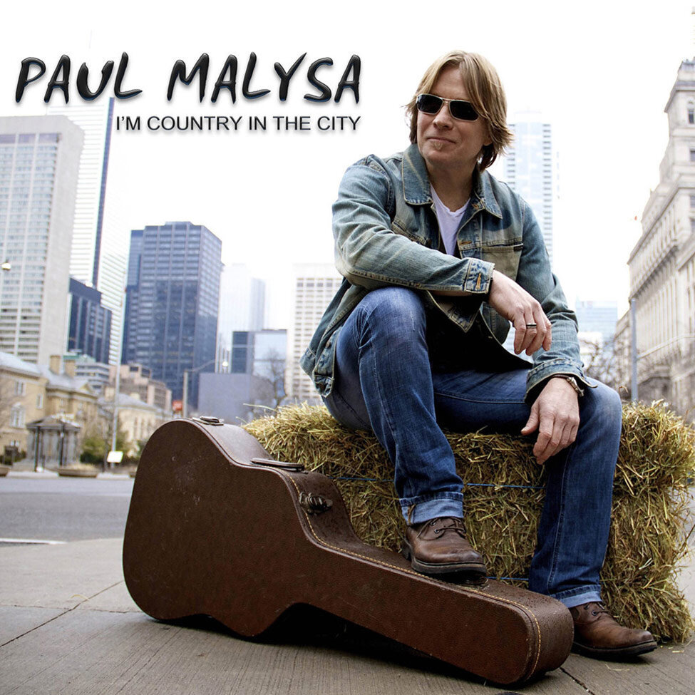 CD Single Cover Title Im Country In The City Artist Paul Malysa sitting on hay bale  on street corner wearing sunglasses foot resting on brown guitar case buildings behind him