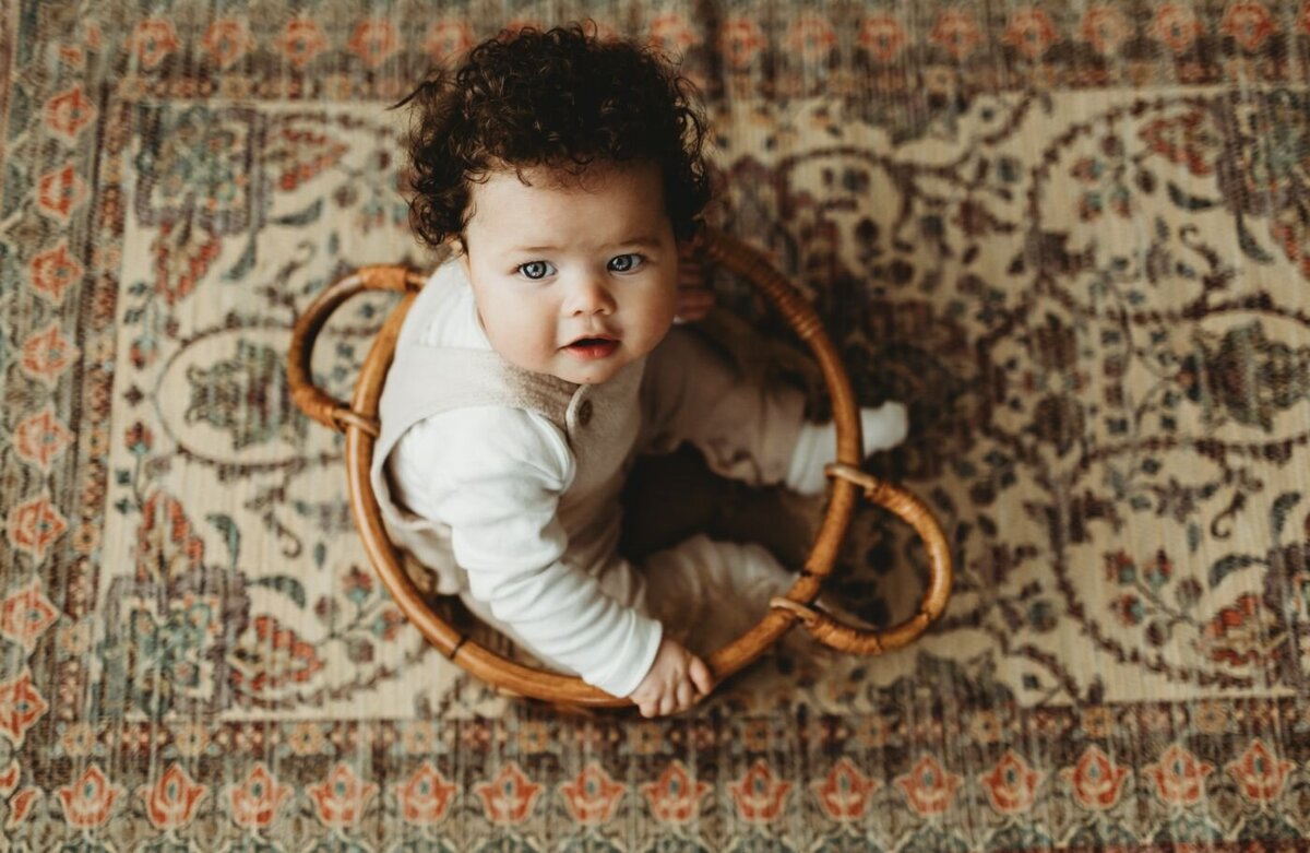 A baby with dark curly hair and bright eyes sits inside a woven basket on a patterned rug.