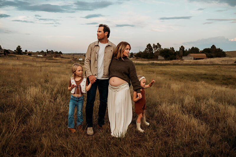 Family of four standing in grassy field on a summer day while mom exposes her growing baby bump