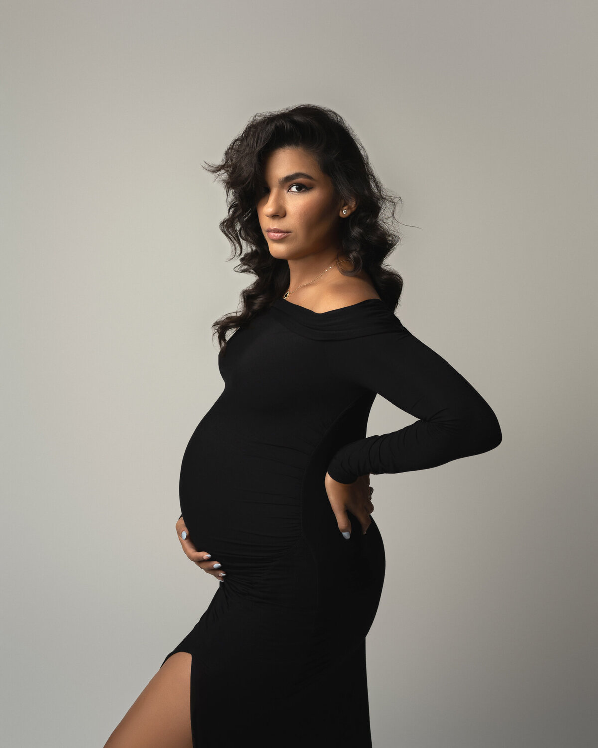 Confident studio maternity portrait of woman in a black gown with hand on hip and soft curls.