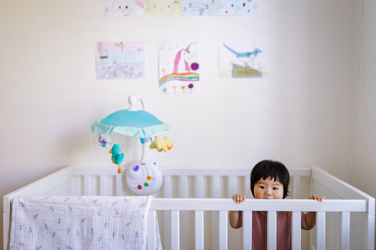 toddler standing in crib with older sister's artwork taped to the wall behind her and she looks at the camera