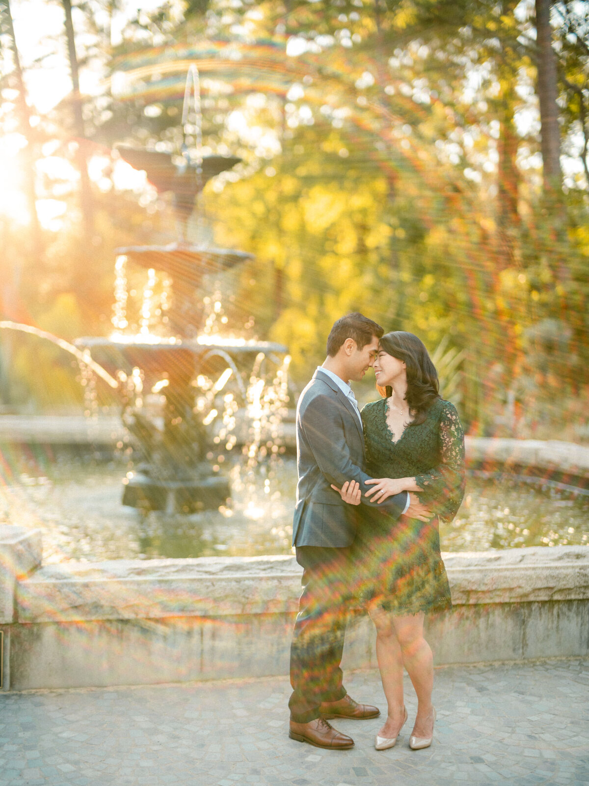 A couple embraced in front of a fountain, the setting sun casting a golden light and water reflections around them.