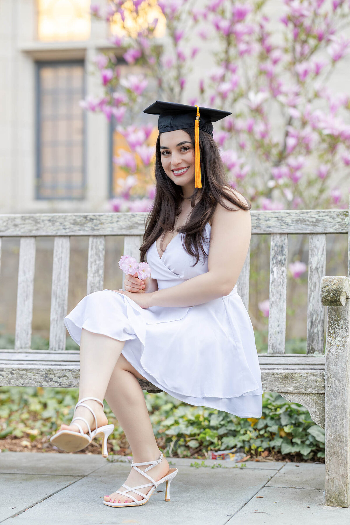 Graduate girl wearing a white dress sits on a bench smiling during her graduate photo session