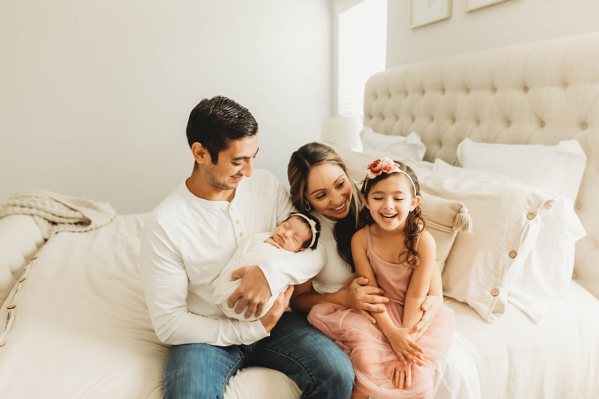 Family of four in a bright bedroom holding their newborn baby.