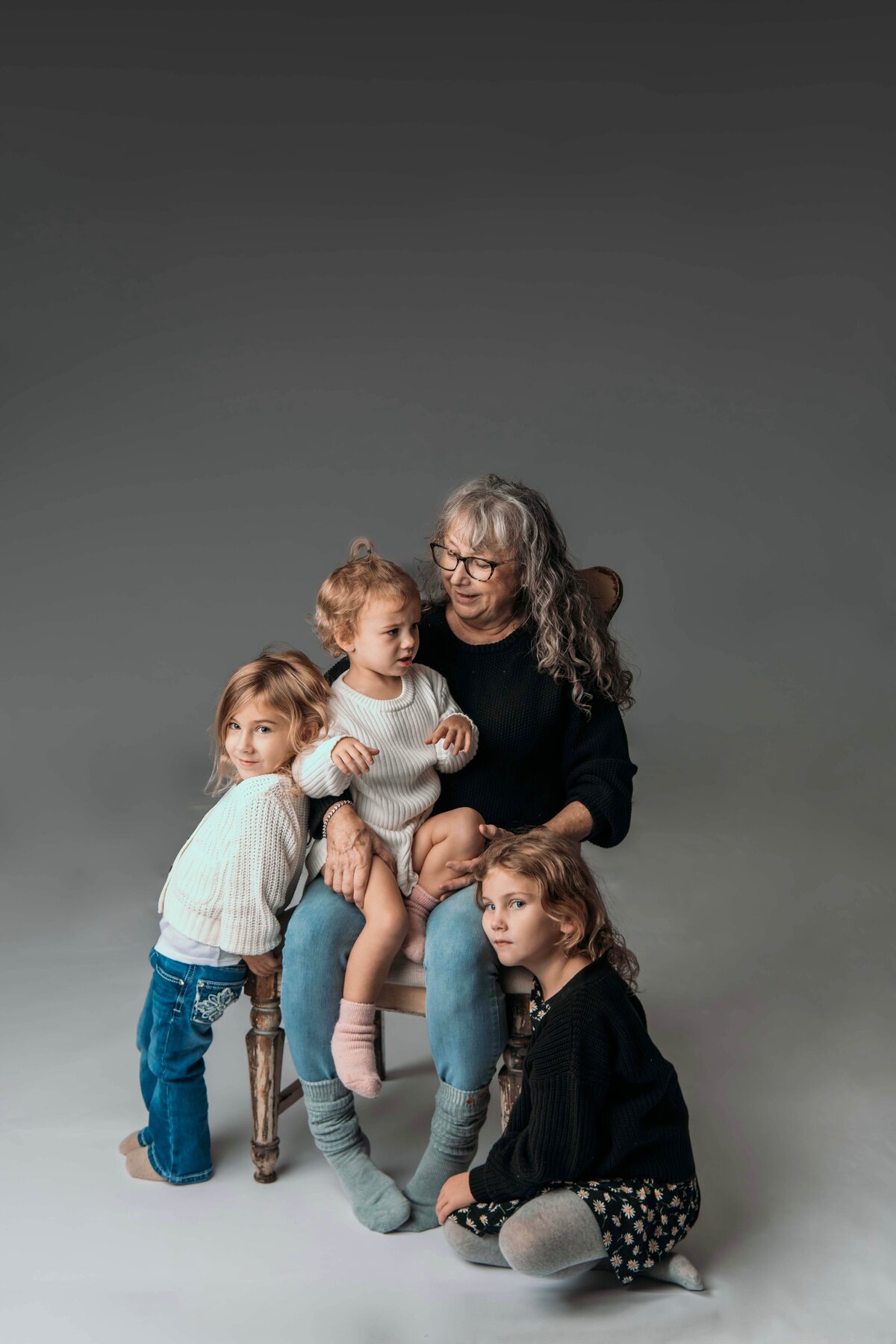 A grandmother sits in a chair with her 2-year-old granddaughter in her lap, while her 4-year-old granddaughter stands on the left, leaning against the chair, and her 6-year-old granddaughter sits on the right, also leaning onto the chair. Each of the three granddaughters is making a different facial expression, showcasing their unique personalities, while the grandmother smiles warmly. The image captures the lively and affectionate family dynamic in Mobile, AL Motherhood Photographer’s studio