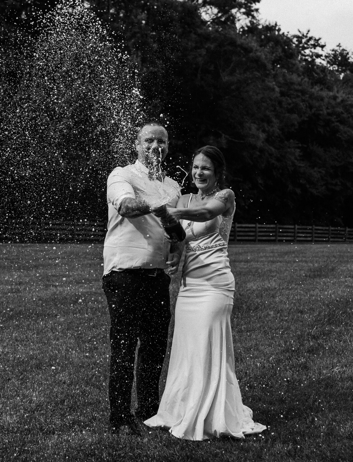 A happy newlywed couple, smiling and laughing, celebrates their marriage by popping champagne in a field at Gentry Farm in Danville, Virginia, capturing a fun and candid wedding moment.
