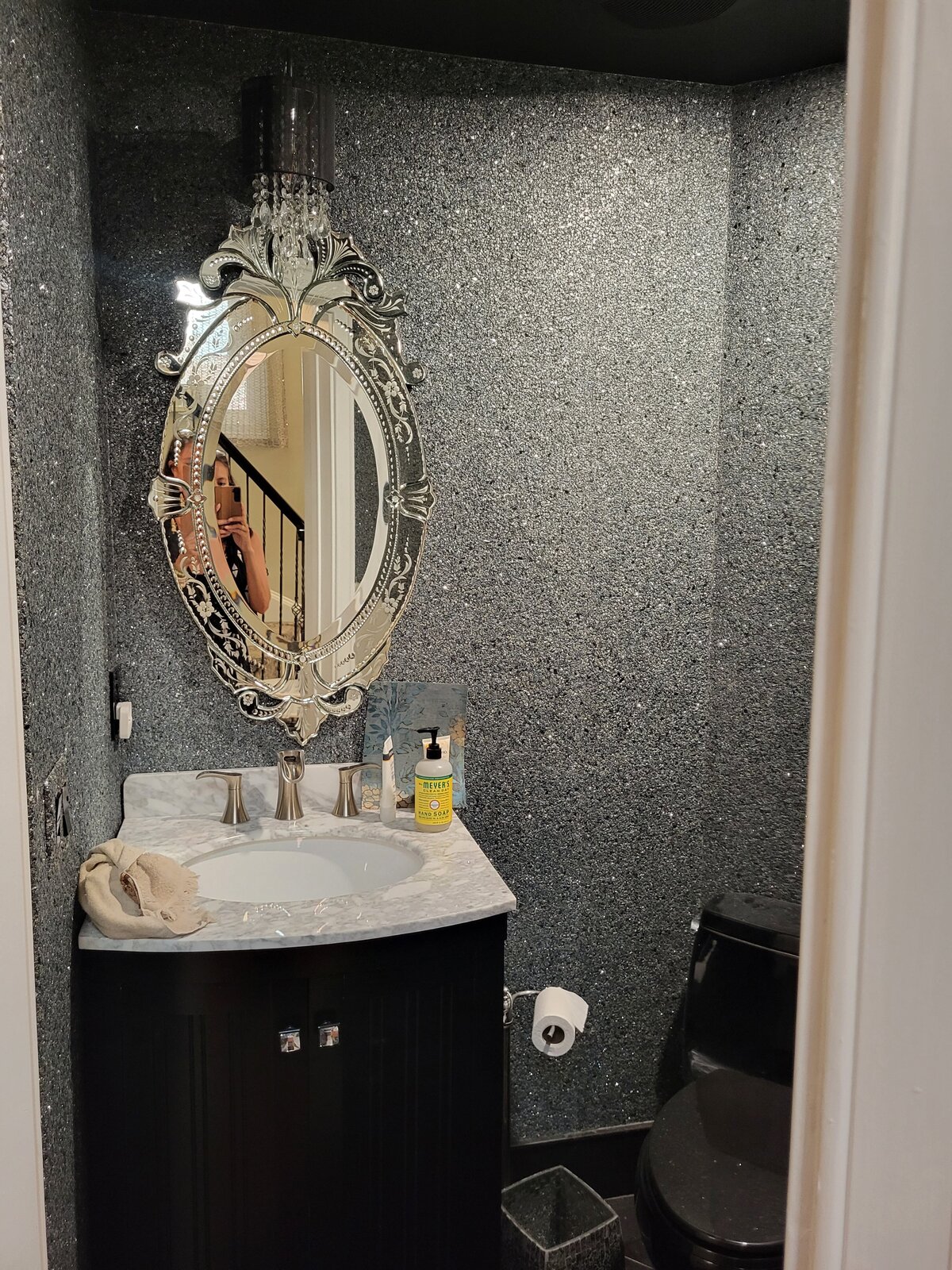 bathroom with mica rock wallpaper and etched mirror over black vanity with marble top and a black tile floor and black toilet with small crystal light