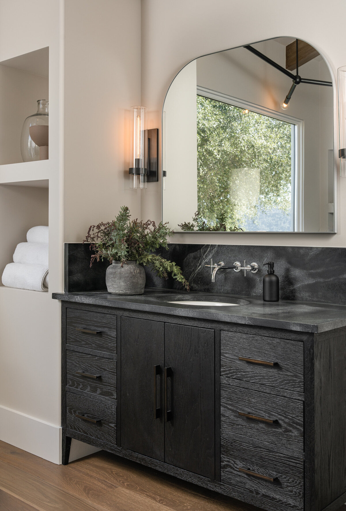 dark wood floors in a bathroom black vanity dark hardware soapstone countertop wall mount faucet arched mirror sconces integrated built-in