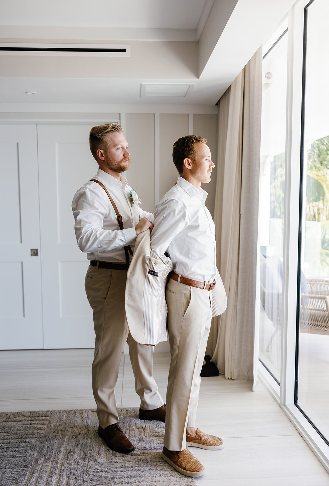 Hunter stands by the window as his groomsman helps him put on his beige suit.