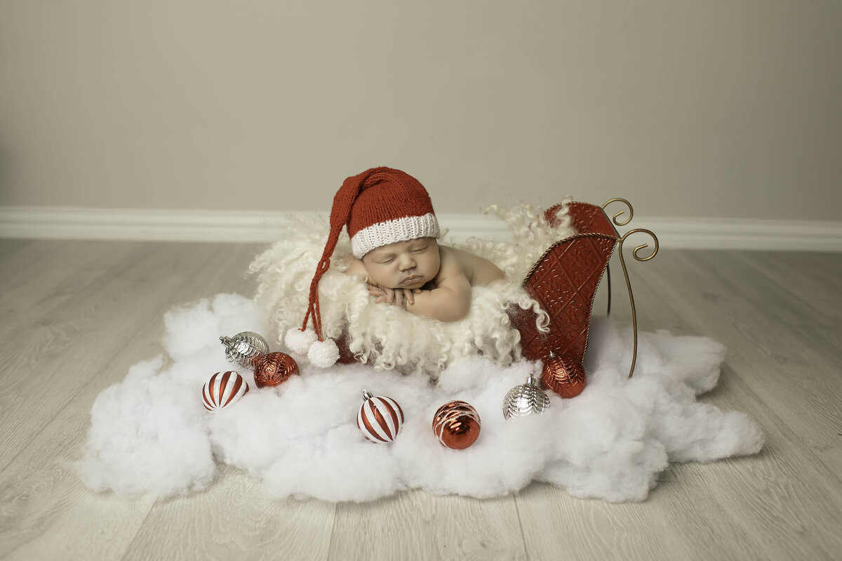 Newborn baby peacefully sleeping in a festive Christmas sleigh, nestled in soft blankets. The holiday-themed setup features warm tones, creating a cozy and magical atmosphere perfect for a newborn's first Christmas