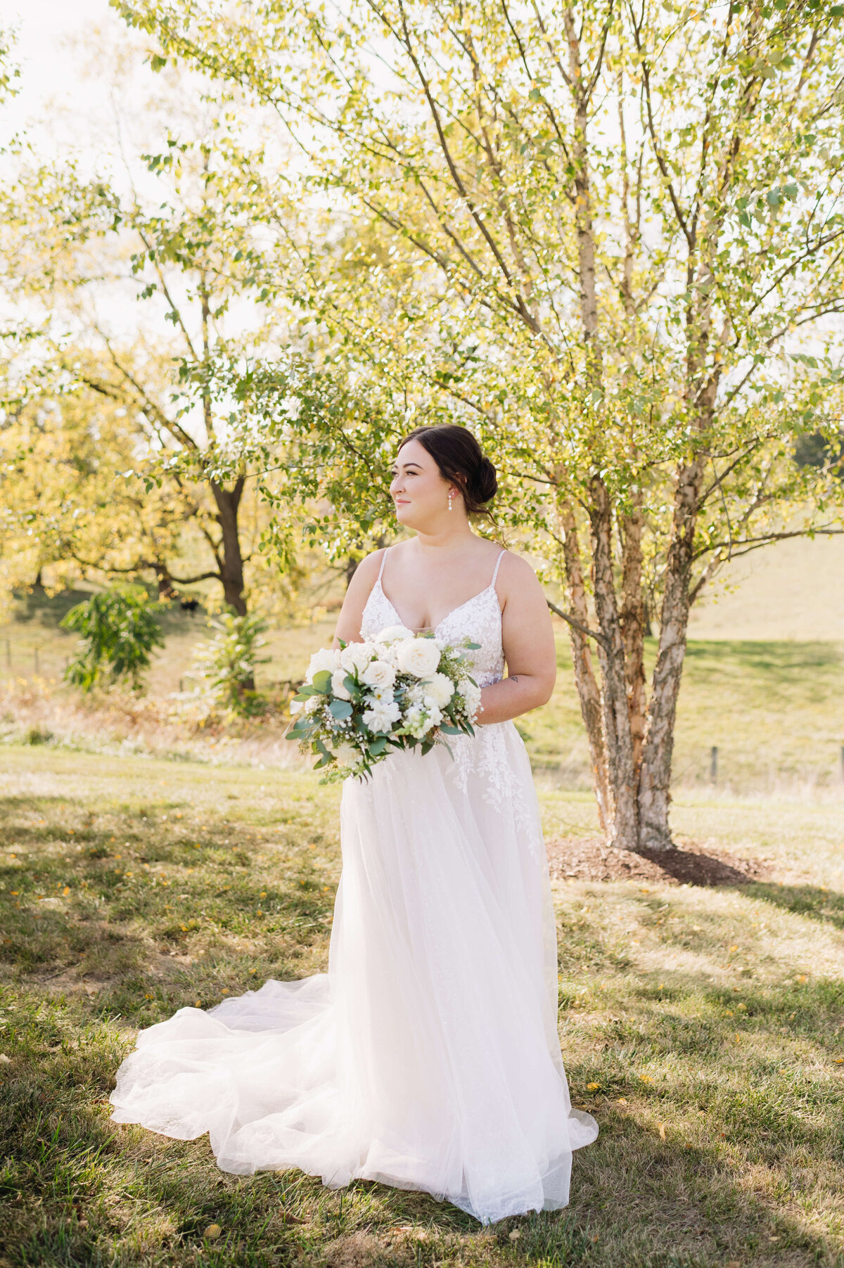 Charlottesville wedding photographer captures bridal photo of bride in a tulle wedding dress holding. awhite floral wedding bouquet with a forest behind her as she smiles over her shoulder