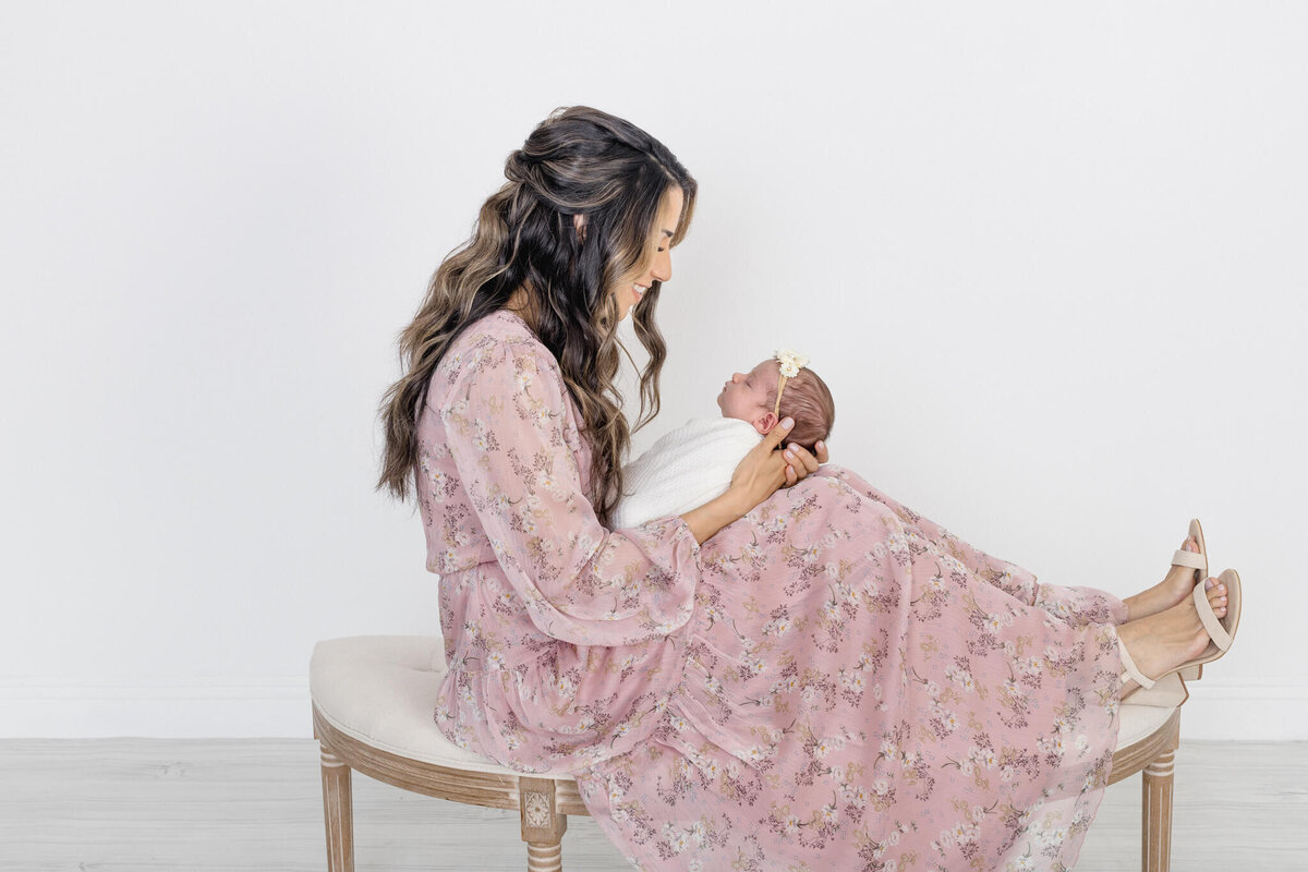 A woman in a pink floral dress sits on a bench, gently holding a swaddled baby with a white bow. They are facing each other, and both are smiling. The background is a plain white wall.