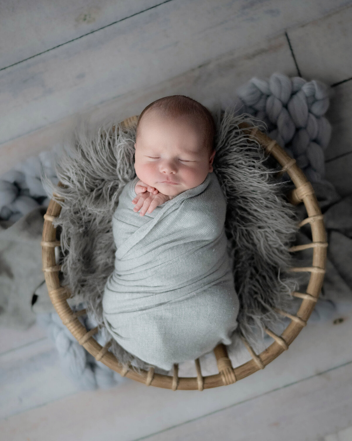 A newborn sleeps in a grey swaddle on fur blanket in a wicker basket