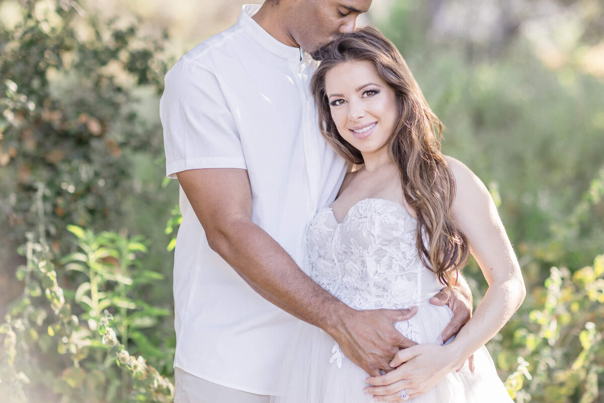A couple stands outdoors surrounded by greenery. The man, wearing a white shirt, gently kisses the womans forehead. The woman, in a strapless white dress, smiles and holds her pregnant belly with both hands.