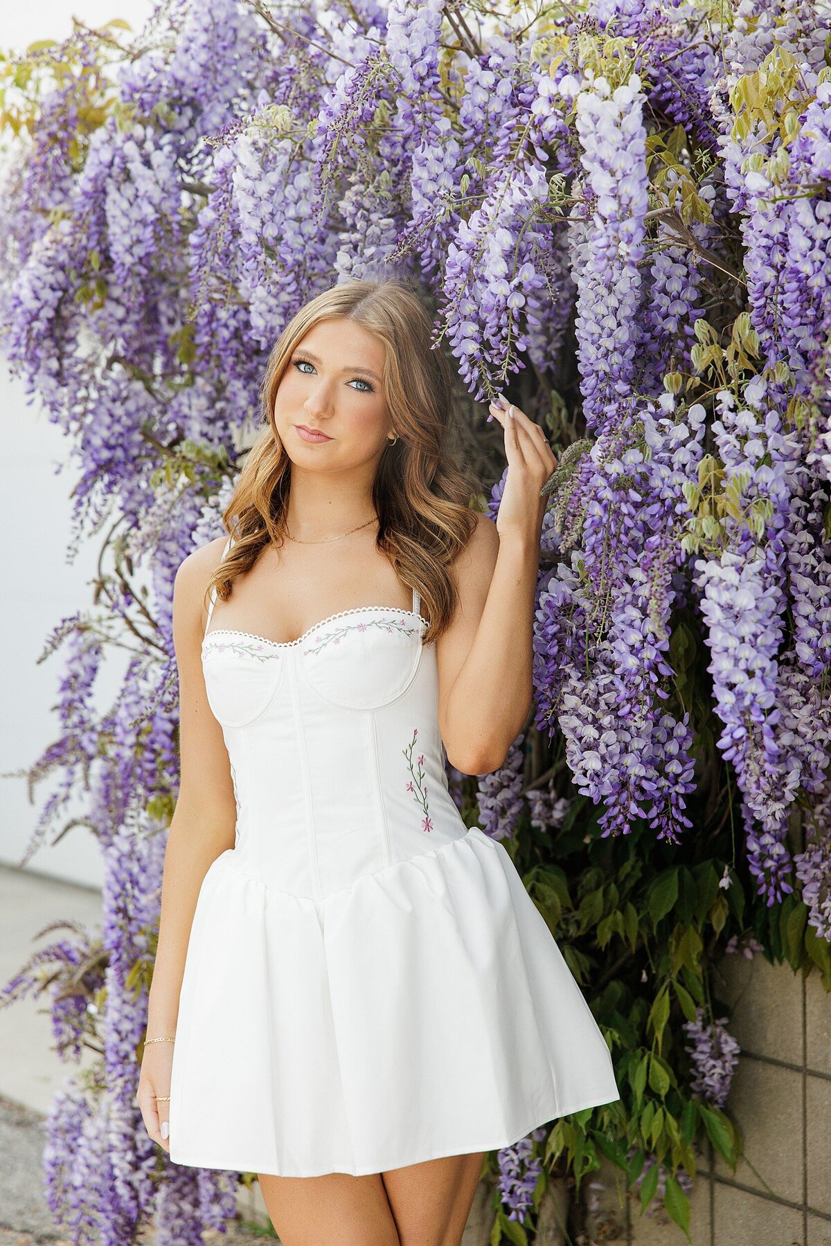 Senior portrait of a teen in a white dress standing in front of blooming purple wisteria, shot in Los Angeles