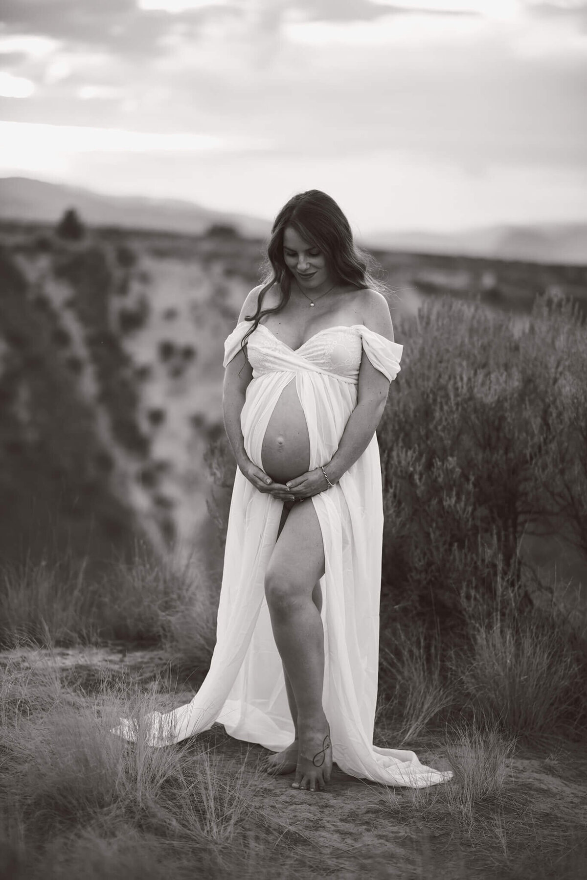 A pregnant mom stands against a beautiful cliff backdrop wearing a white flowy dress. She looks down at her belly while caressing it. Taken by Brittany Danielle