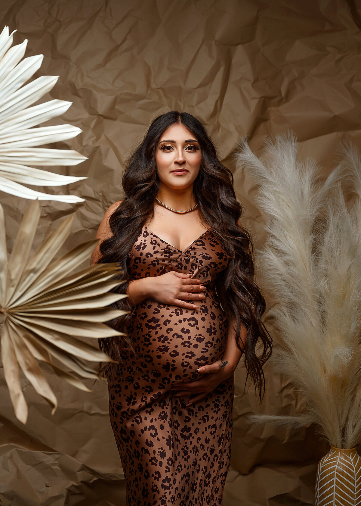 Gorgeous pregnant mom in a  cheetah print dress against brown crinkled paper background with dried palm leaves and pampas grass around her