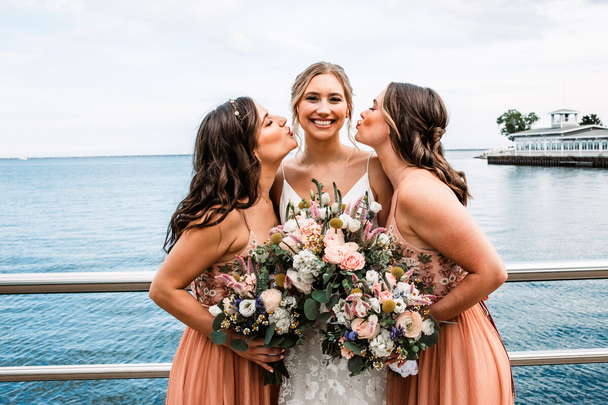 Two bridesmaids pretend to kiss the cheeks of the bride by the water