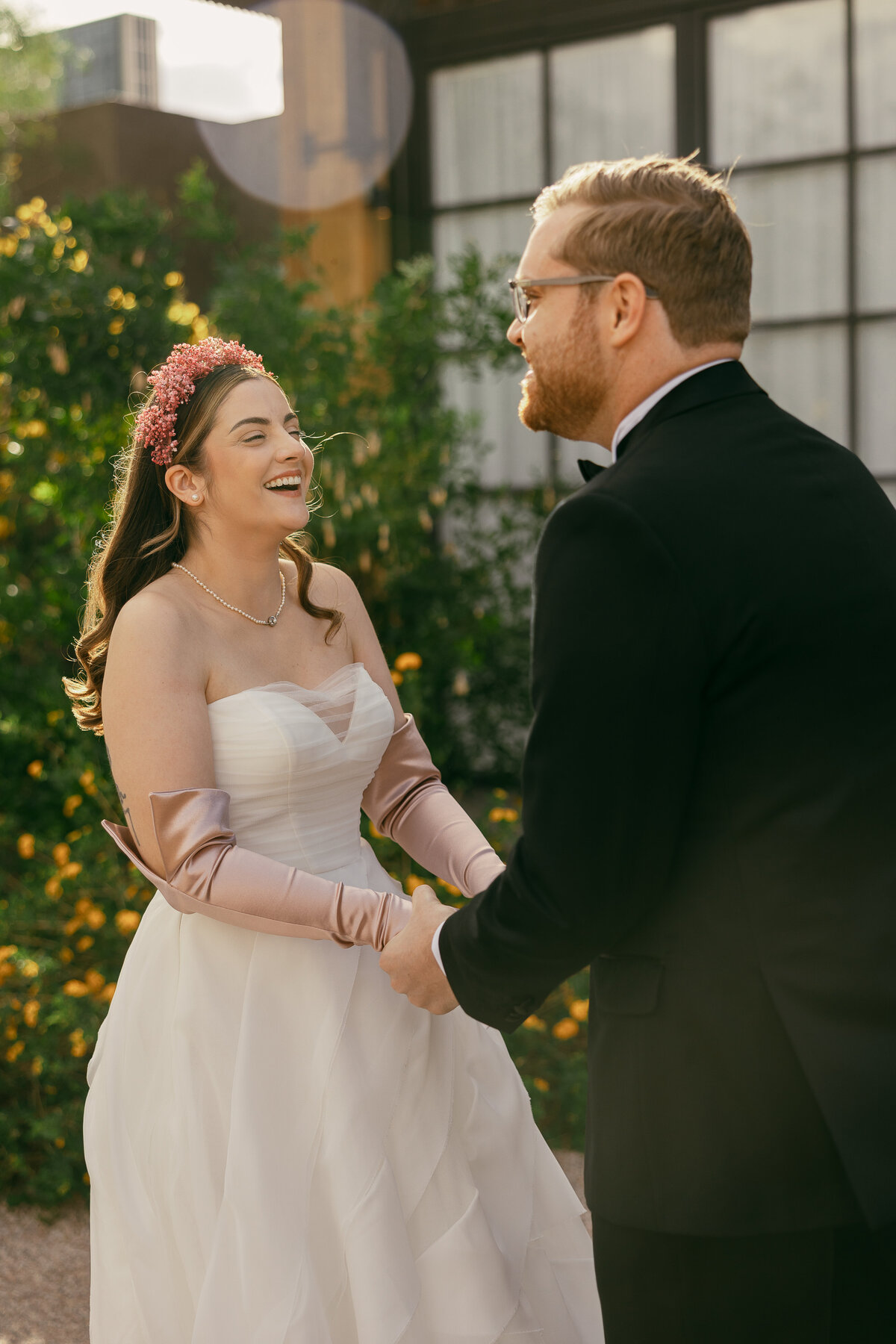 a bride in a white dress with pink gloves and floral headband laughs as she holds the hands of her groom