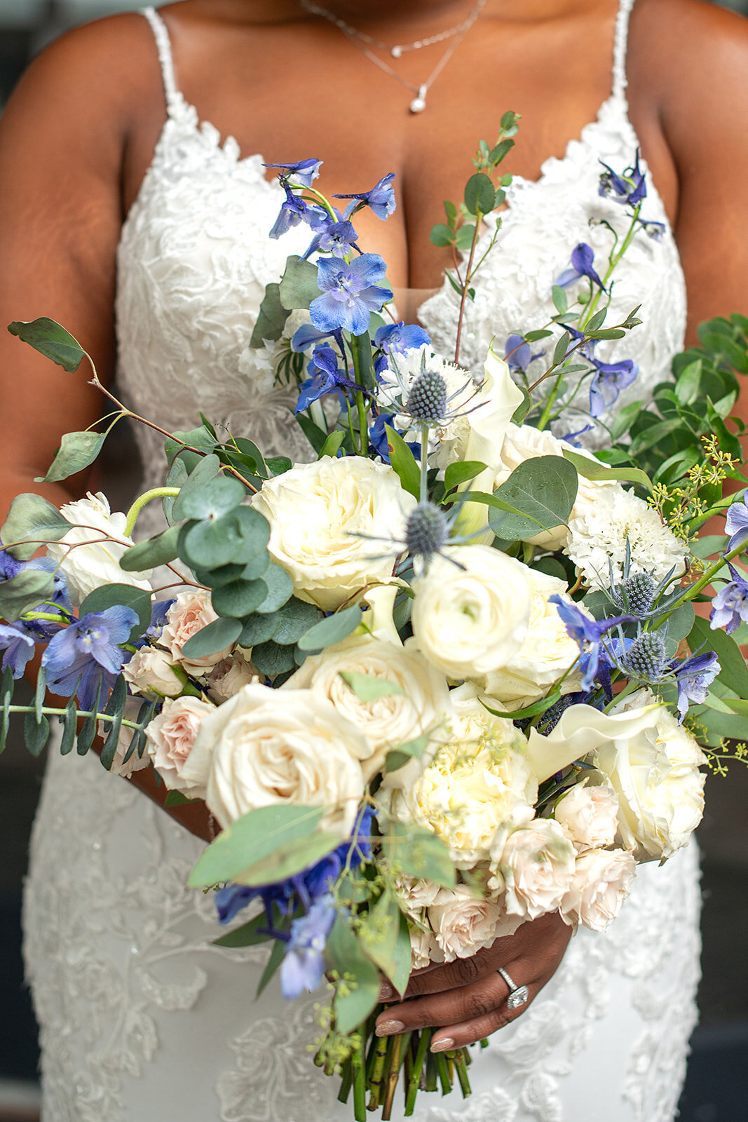 Bridal bouquet featuring white roses, blue delphiniums, and greenery, complementing an elegant lace gown.