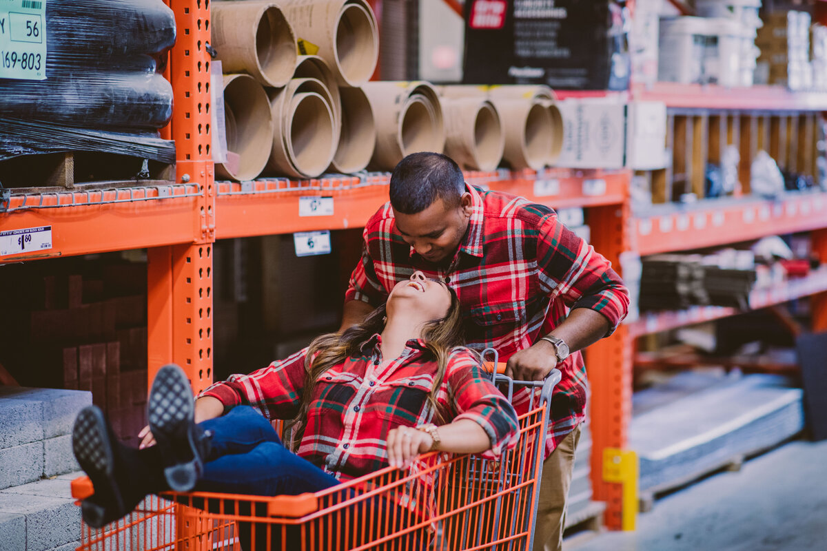 A bride sitting inside a shopping cart inside the home depot is looking up at the groom steering the cart wearing matching plaid outfits