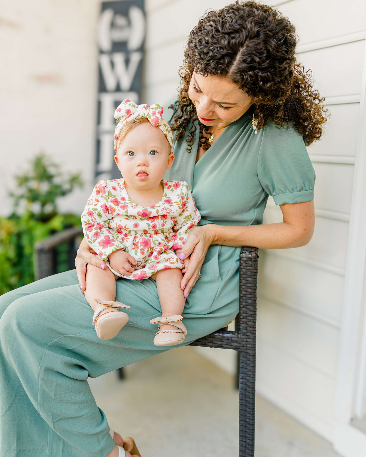 A woman with curly hair sits on an outdoor chair, smiling at a baby in a floral dress and matching headband. Captured by a Fayetteville NC family photographer, the scene features part of a white house and lush greenery in the background.