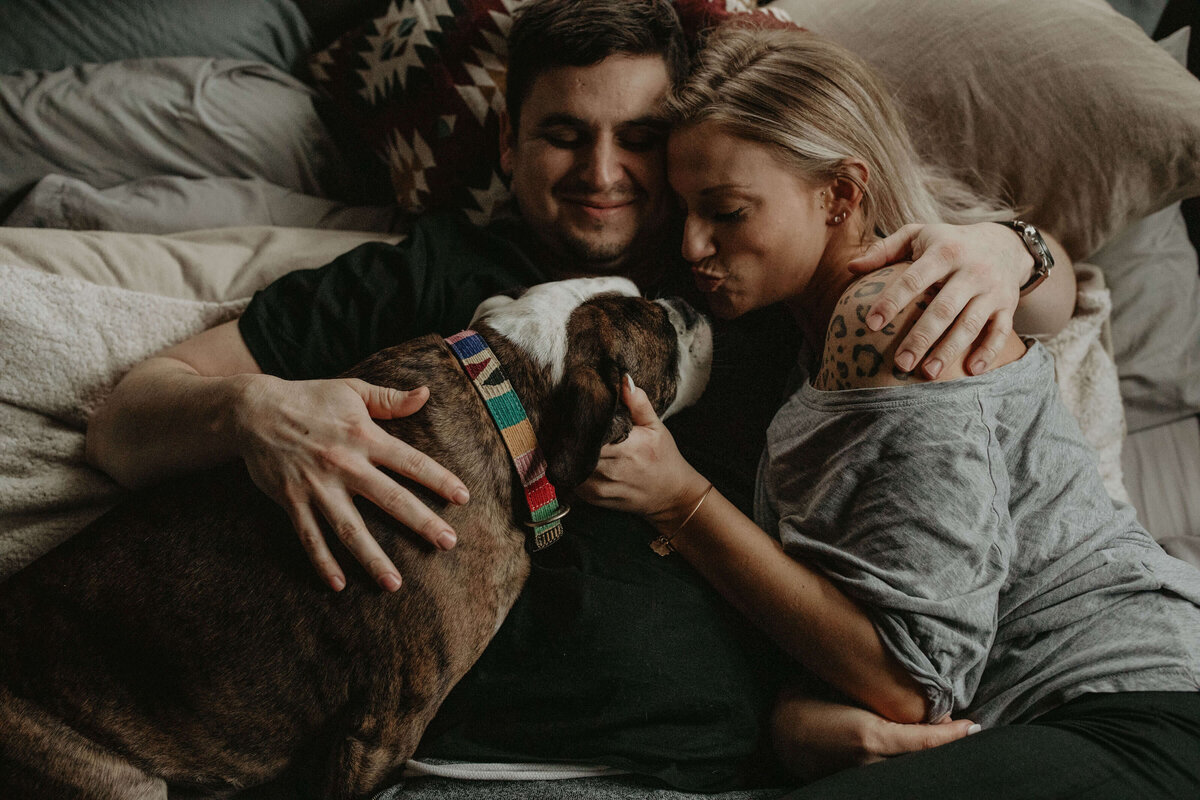 A couple lies in bed, hugging their dog who is nestled between them, all looking content and relaxed.