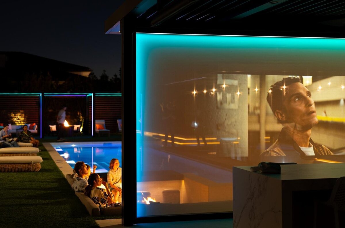 StruXure pergola with TraX system; the TraX LEDs are illuminated teal and there is a movie with Matthew McConaughey being projected onto the retractable screen. 