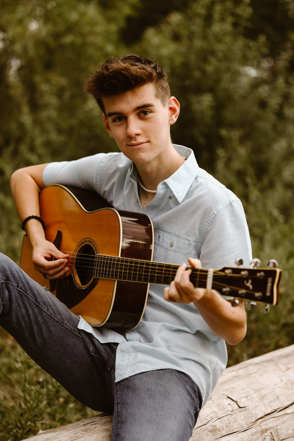 A senior boy sits on a log playing an acoustic guitar, wearing a light blue shirt and jeans, surrounded by green foliage.