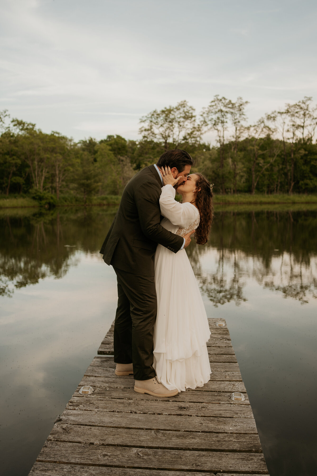 A bride and groom share a romantic kiss on a wooden dock by a serene lake at Blenheim Hill Farm. The bride, wearing a flowing white dress with long sleeves, holds the groom's face gently. The groom, dressed in a dark suit and light-colored shoes, embraces the bride. The tranquil lake and lush green trees in the background are perfectly reflected in the water.