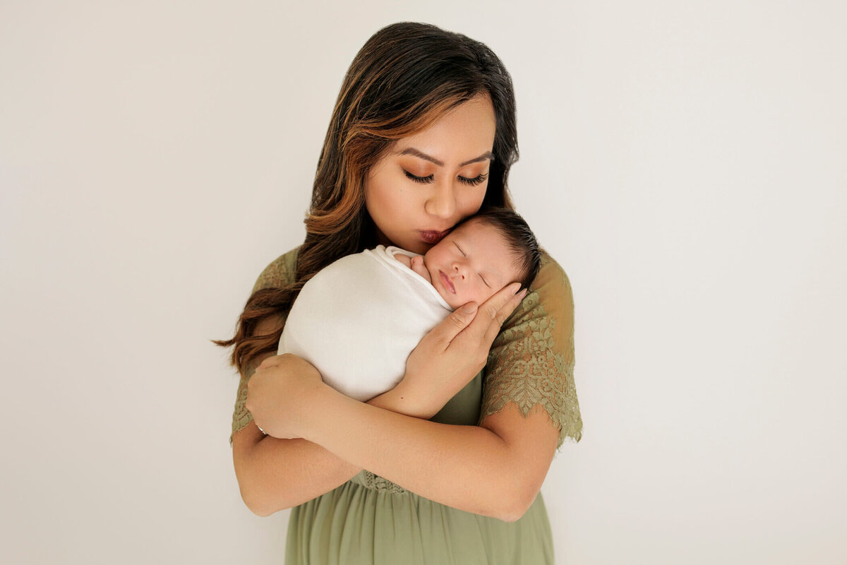 Mother lovingly holding and kissing her newborn baby wrapped in a white blanket.