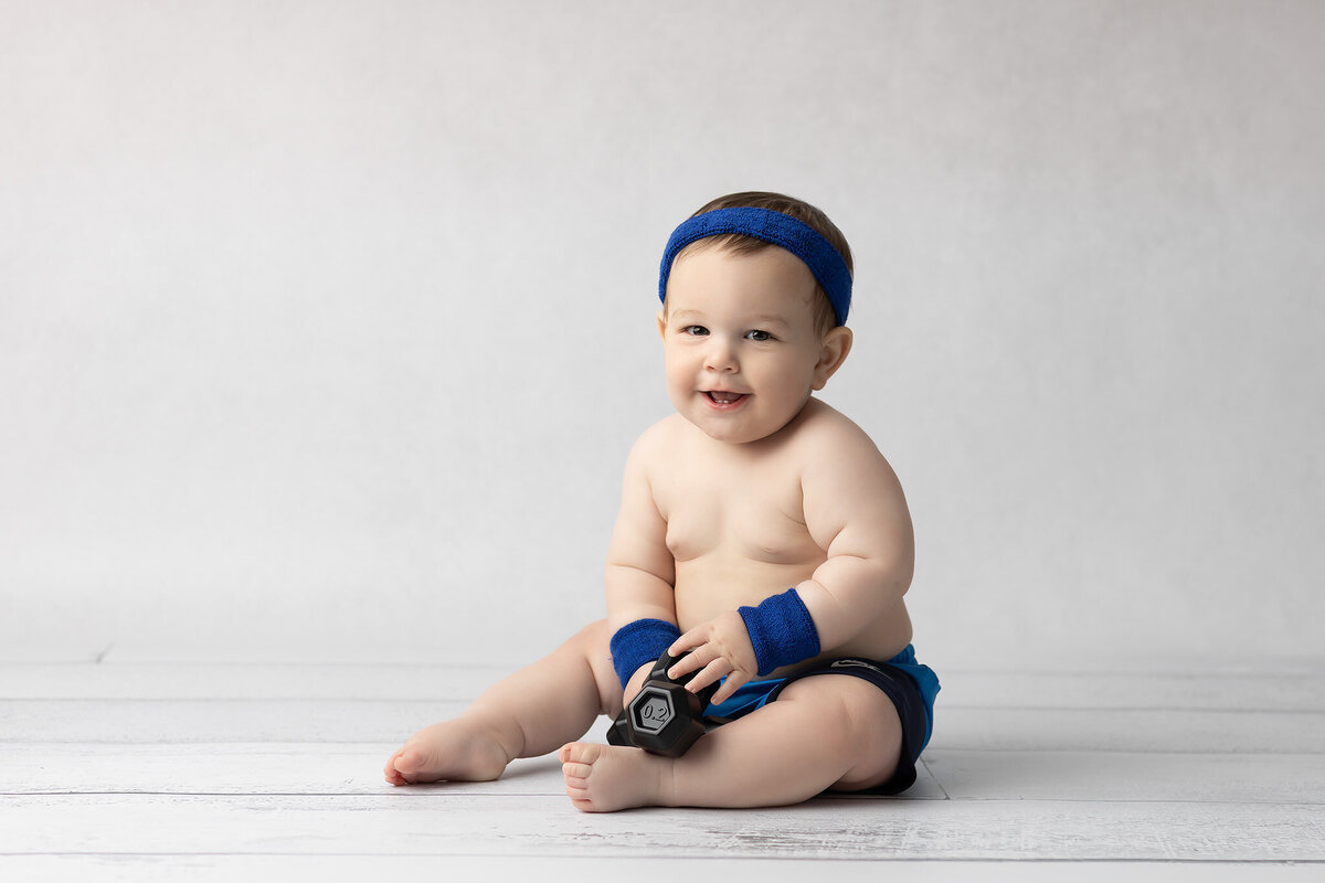 Chubby baby in blue workout gear and sweatbands holds a toy dumbbell while sitting on a white wood floor, captured by a DFW milestone photographer.