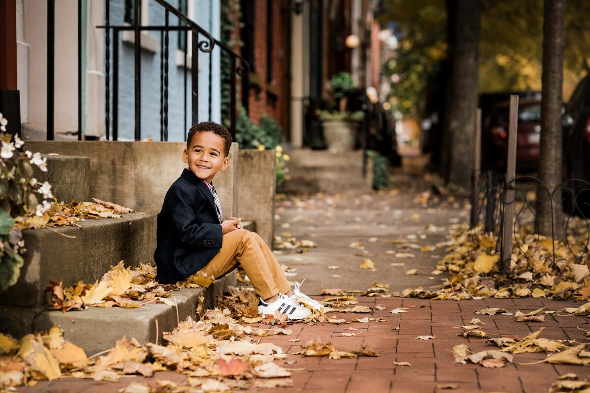 A Pittsburgh family photographer captures a young boy sitting on a step amidst autumn leaves, smiling at the camera.
