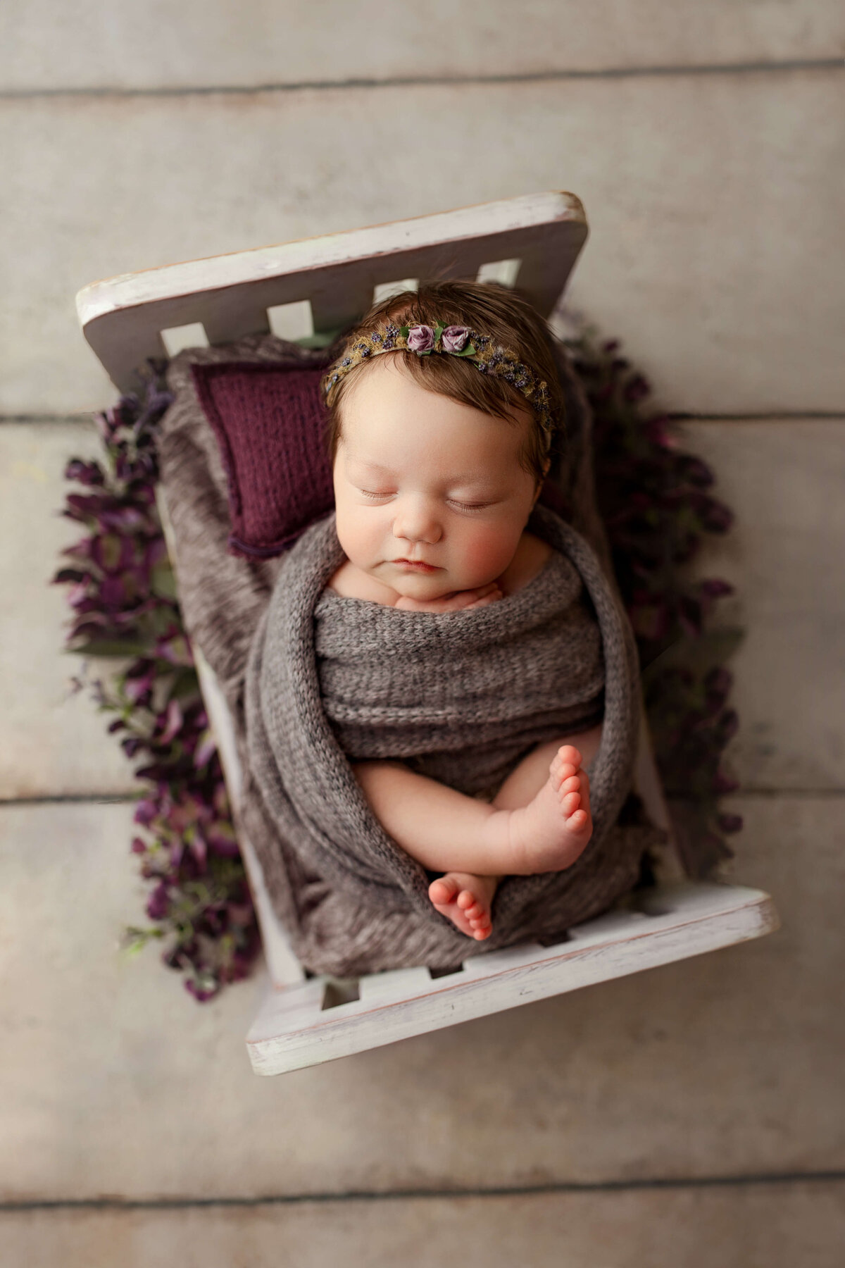 newborn baby in a purple wrap laying    on a white wooden bed with flowers around her