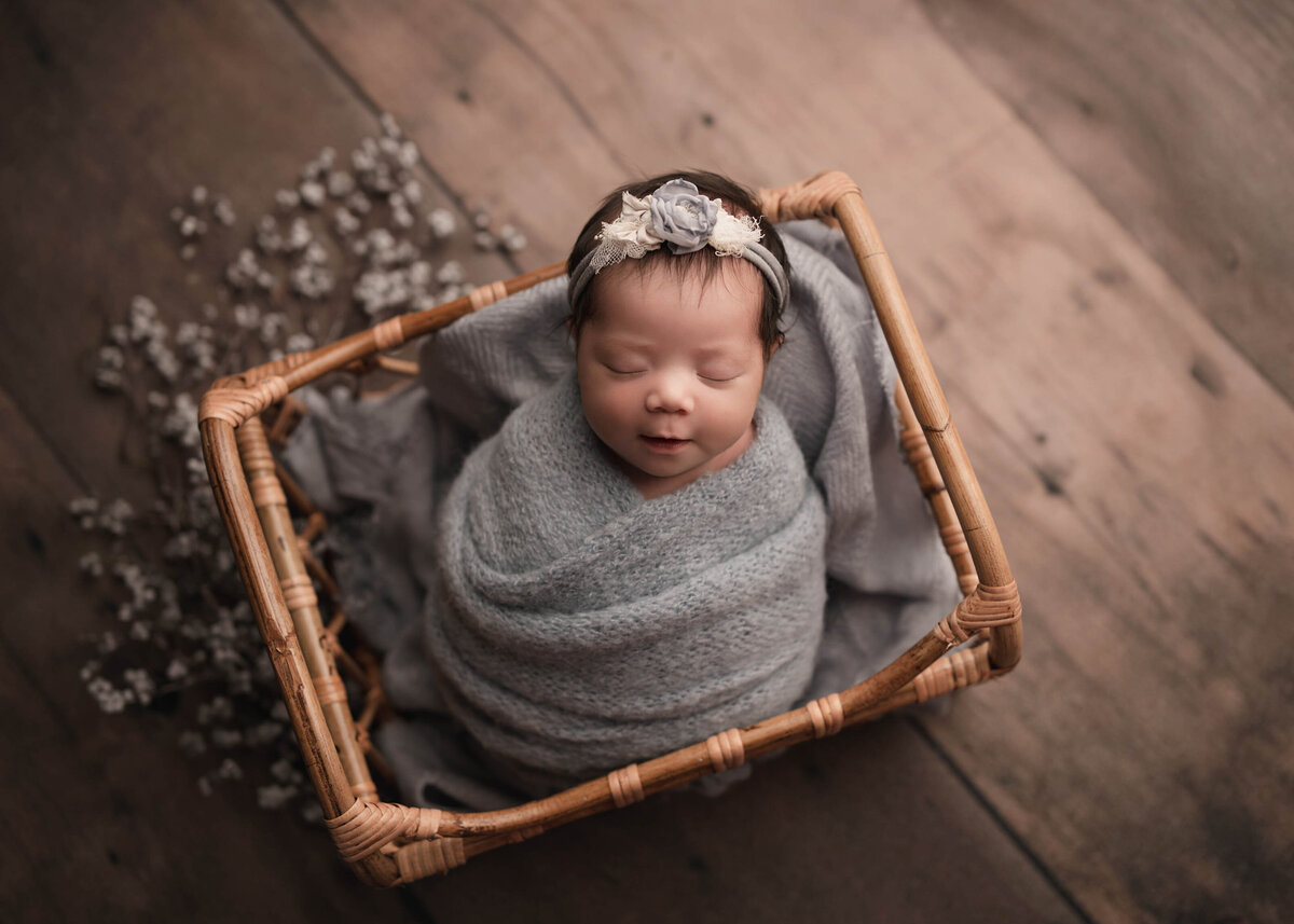 Aerial image. Baby girl wrapped in grey knit swaddle  for her newborn photoshoot. She is laying in a basket and wearing a grey and cream headband. Captured by best Menifee newborn photographer Bonny Lynn Photography