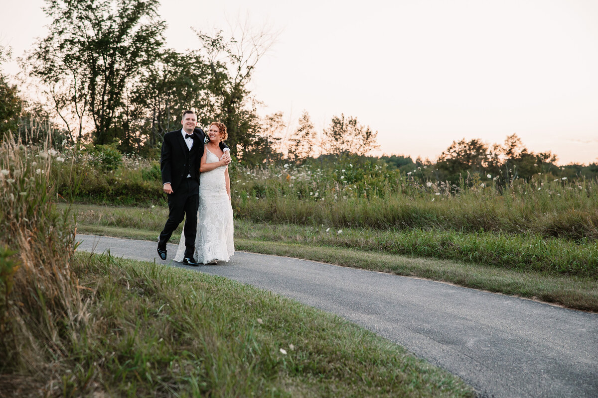 Newlyweds walk with groom's arm around his bride on a sidewalk at sunset