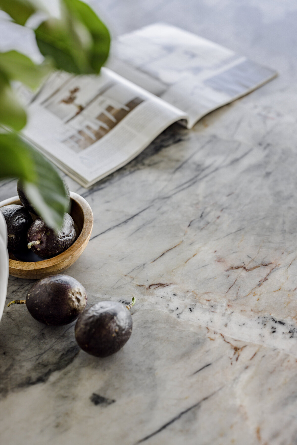 A lush plant complements the vibrant passion fruits on a marble table, with an open magazine inviting inspiration from a Newcastle Kitchen and Bathroom Designer in the background.