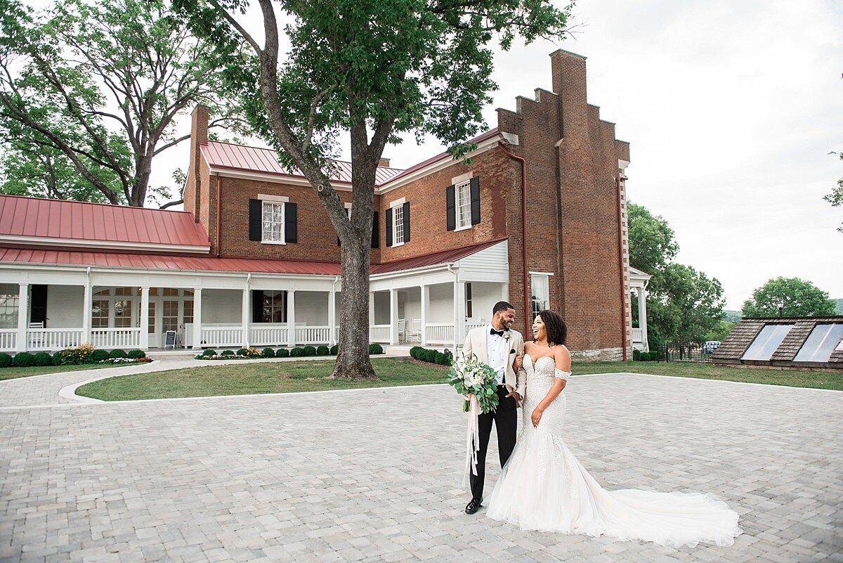 The African American bride and groom, Jasmine Sweet and Alex Sweet, share a private moment in the courtyard of Ravenswood Mansion. The groom i swearing a tuxedo with a white jacket and the bride is wearing an off the shoulder lace mermaid style wedding dress with a long train for their summer wedding.