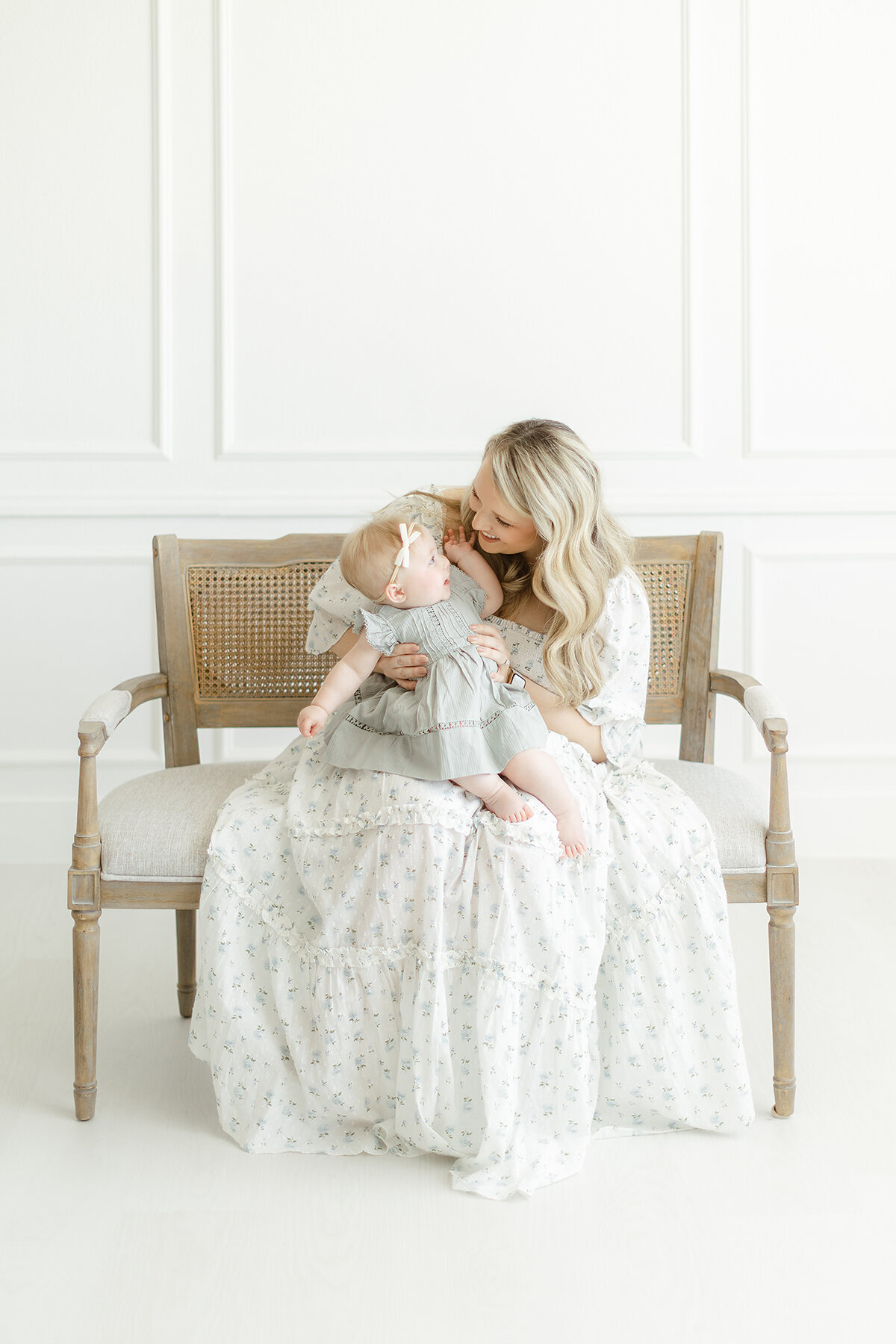 A mommy and me session taken in a Dallas photography studio of a mother sitting on a bench playing with her baby girl.