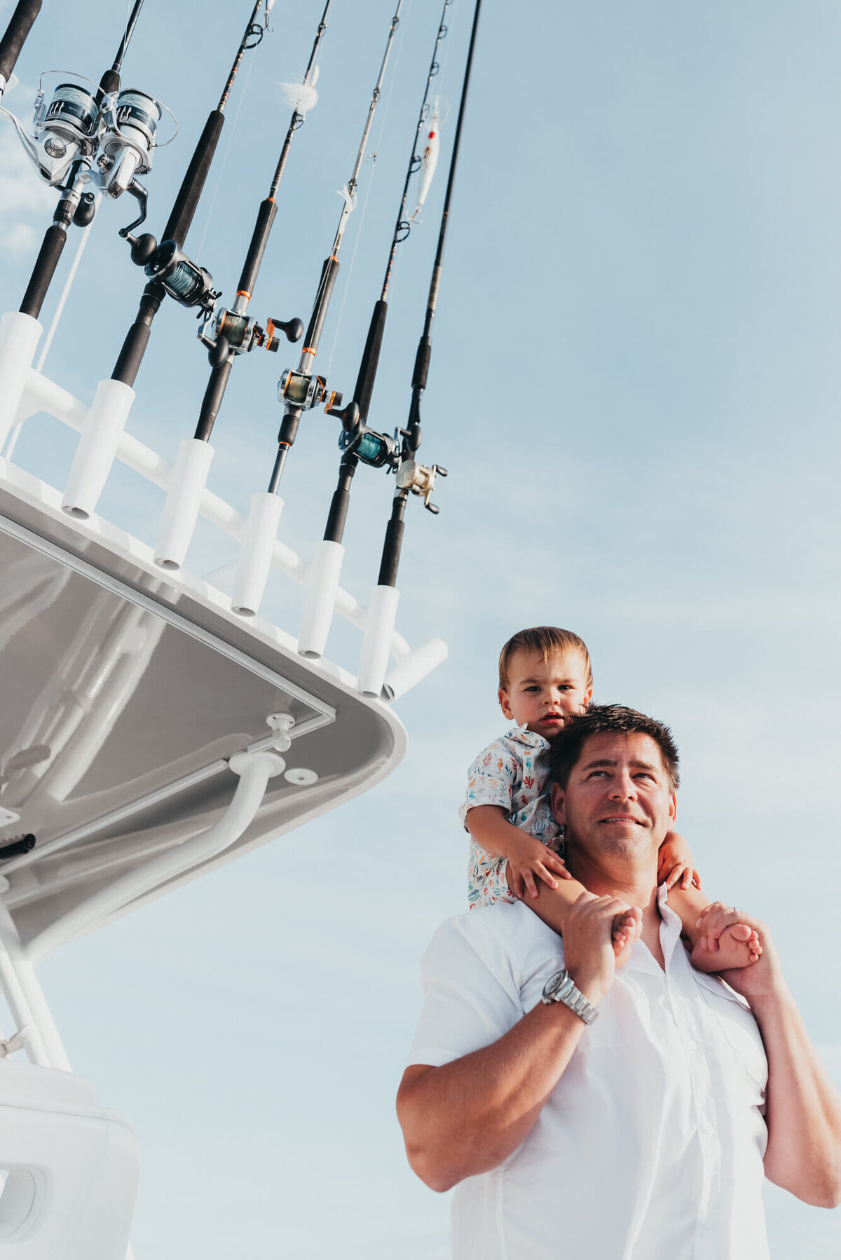 A father holds his son on his shoulders on their boat, you can see all of their deep sea fishing rods in the background
