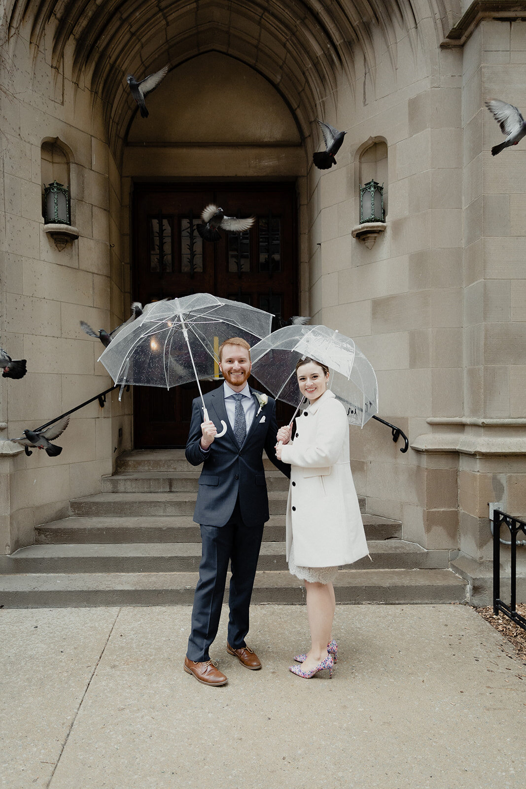 Just Married photo session couple stands with their umbrellas in front of an old Chicago church and pigeons fly around them.