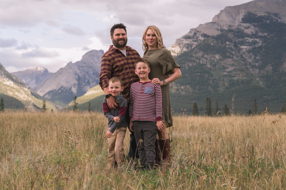Family of four stands smiling with dramatic mountain range behind them in Canmore.