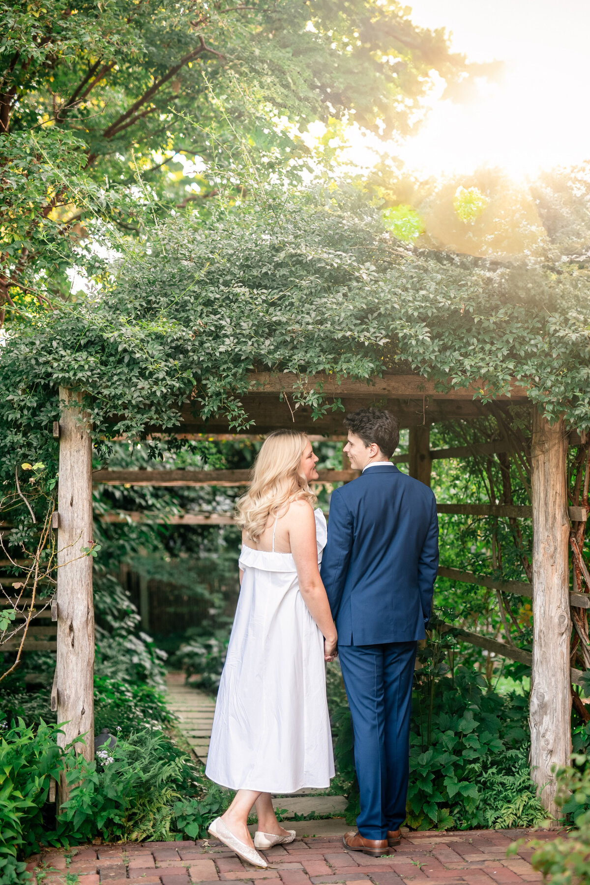 A couple stands hand in hand under a wooden archway in a garden. The woman, in a white dress, and the man, in a blue suit, look at each other. Sunlight filters through the trees, casting a warm glow over the scene.