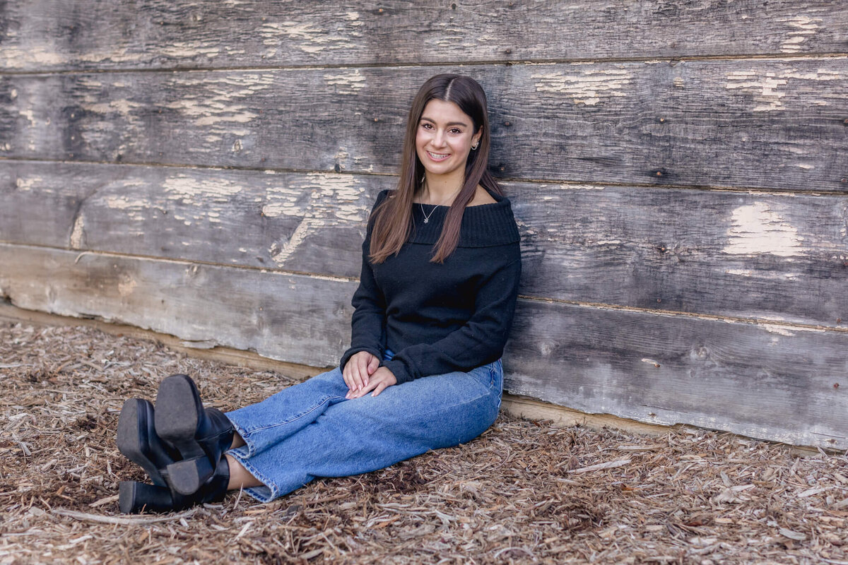 A young brunette woman sitting in wood chips with her feet crossing at the ankles, smiling at the camera and leaning against a grey and beige wooden wall wearing jeans and a black sweater.