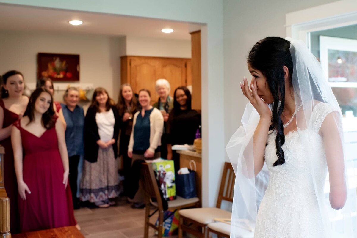a bride wipes tears away as a dozen people see her in her wedding dress for the first time