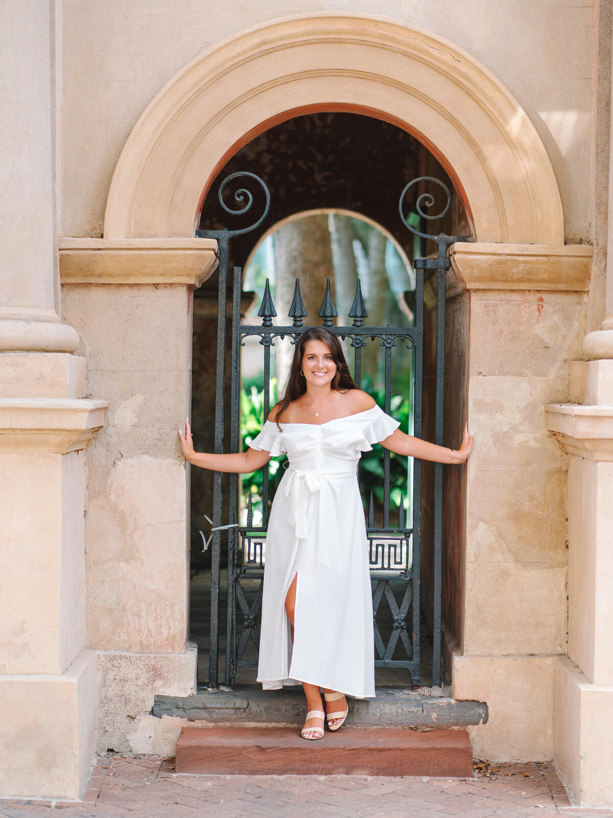 Senior girl posing in white dress under arched stone entryway in historic College of Charleston 