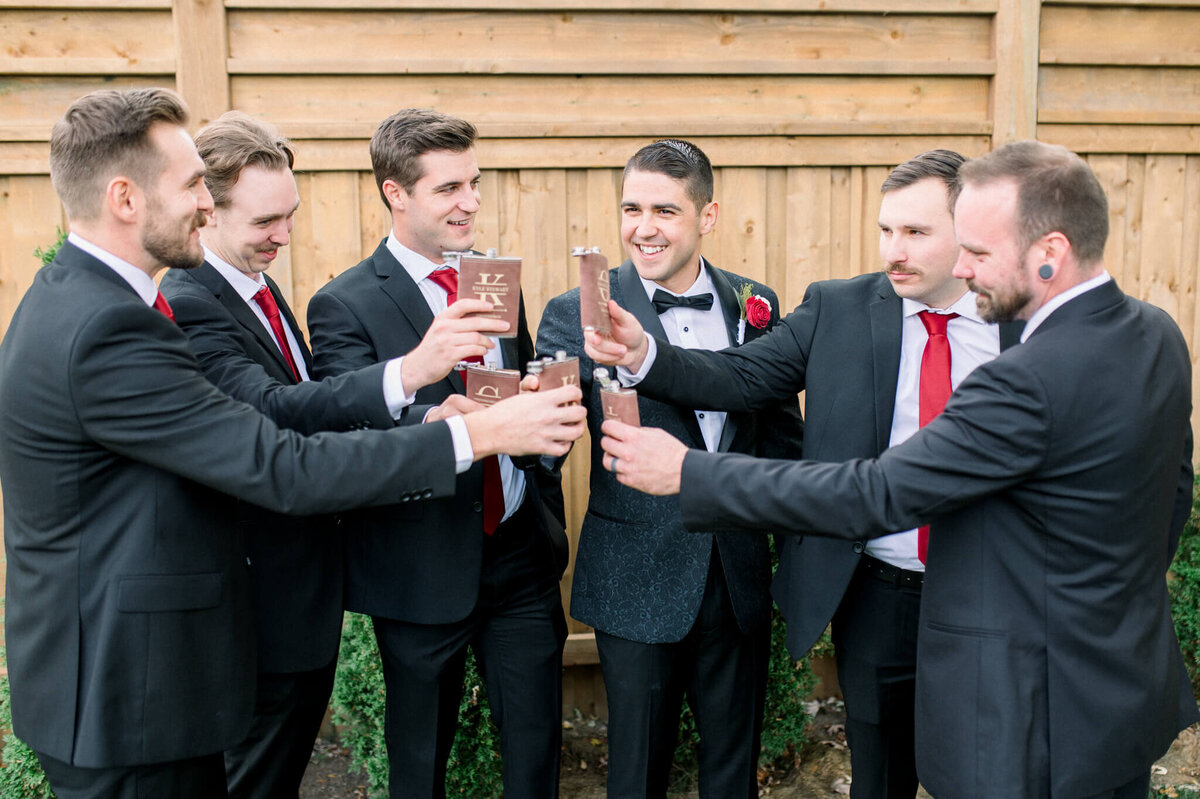 Groomsmen gather in a half circle and cheers with their flasks. Captured by Niagara wedding photographer