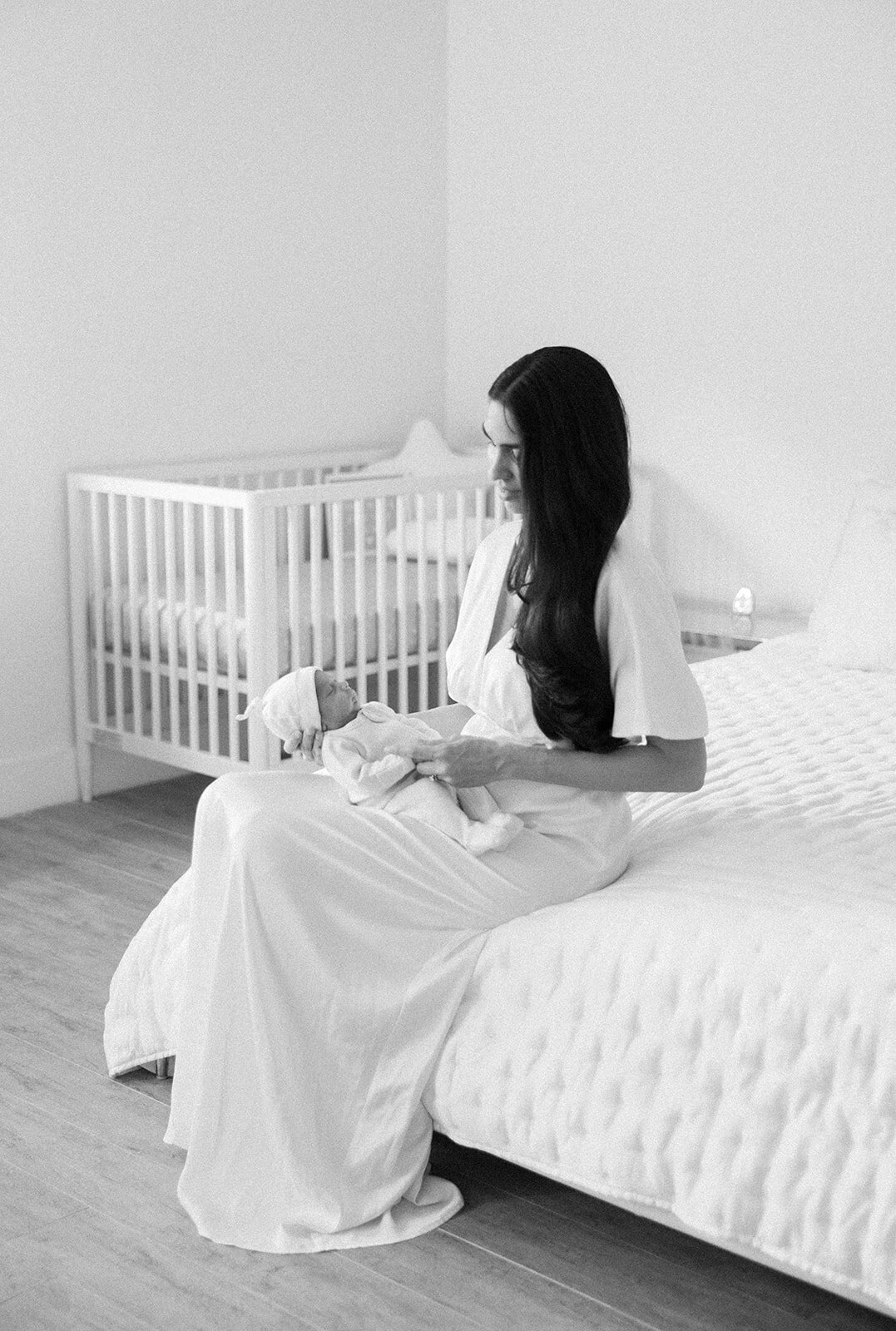 A mother in a flowing white gown cradles her newborn in a softly lit nursery during a black-and-white Florida Keys family session.