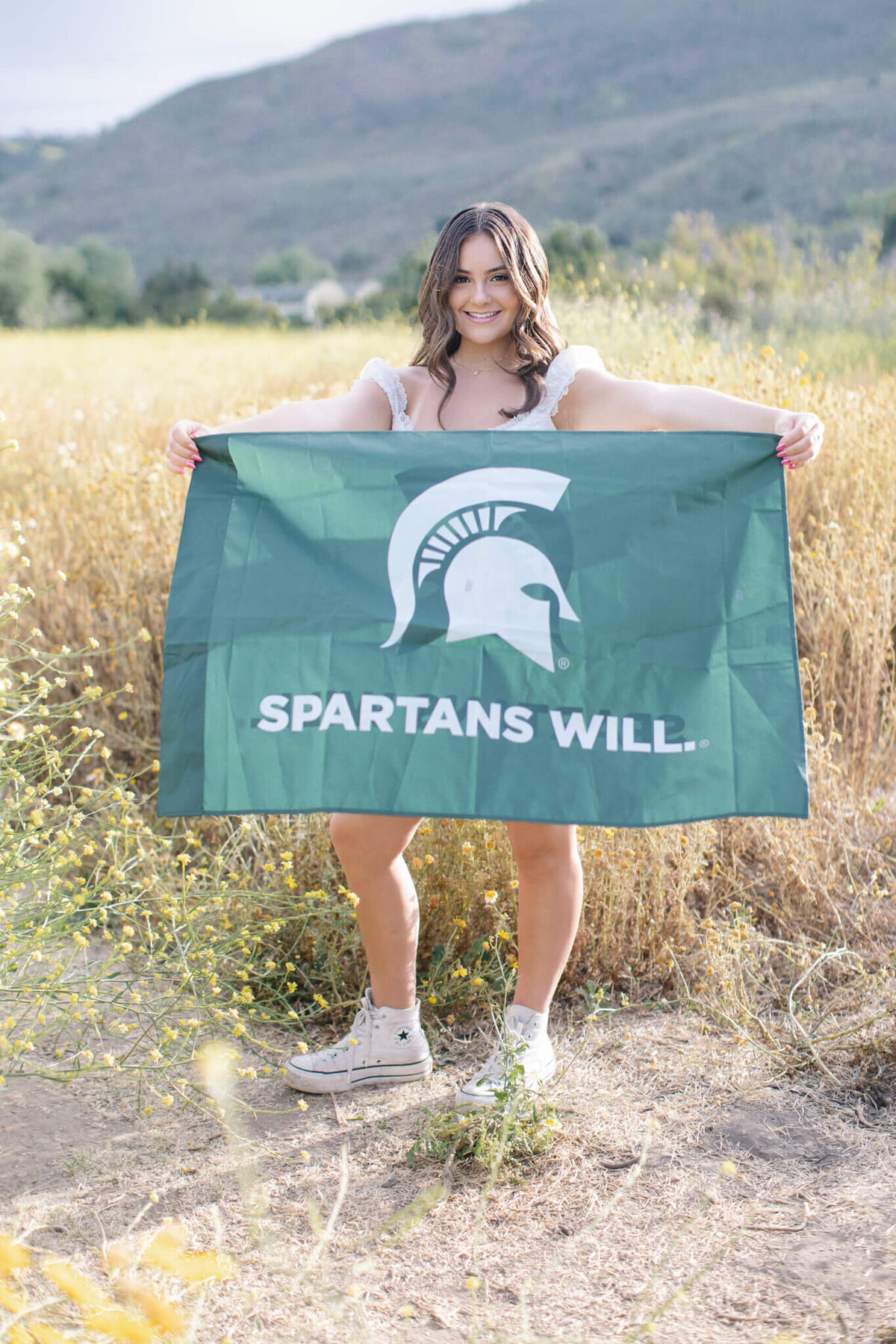 A person stands in a field holding a green flag with a white Spartan helmet and the words SPARTANS WILL. The background features rolling hills and greenery under a clear sky.