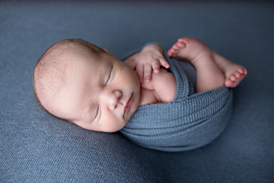newborn boy posed on a denim blue fabric and wrapped in a matching swaddle.  The baby is posed on his back with his hands and legs peeking out of the swaddle.
